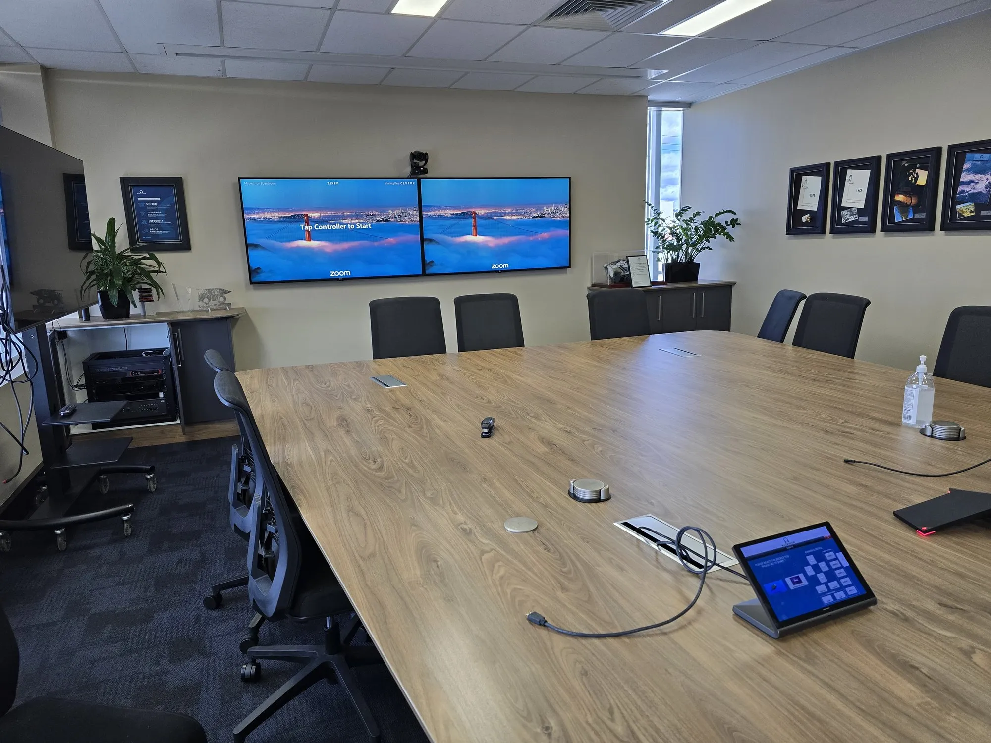A boardroom table with a touch controller in the foreground and dual displays beyond