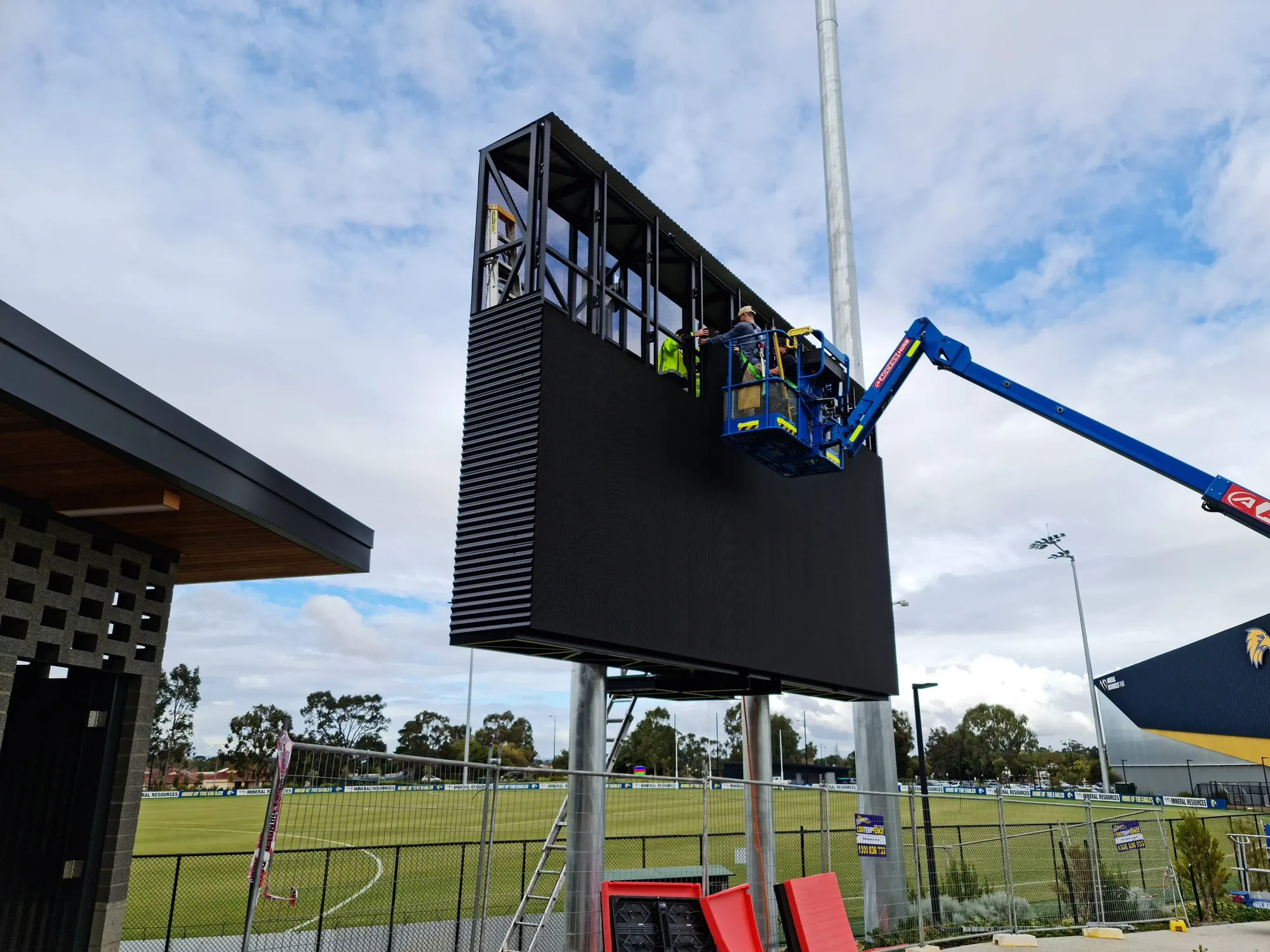 Technicians in a boom lift fitting the final panels to the West Coast Eagles scoreboard, cabinets staged on the ground