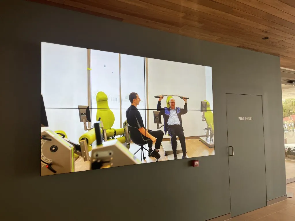 A four-panel video wall in a Brightwater Care Group community space showing residents exercising in the on-site gym