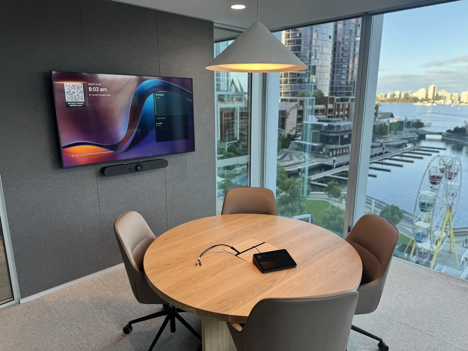 A four-person meeting room at Multiplex's Nine The Esplanade office in Perth — a commercial display with an all-in-one video bar beneath it, a touch controller on the round table and the Swan River through the glass