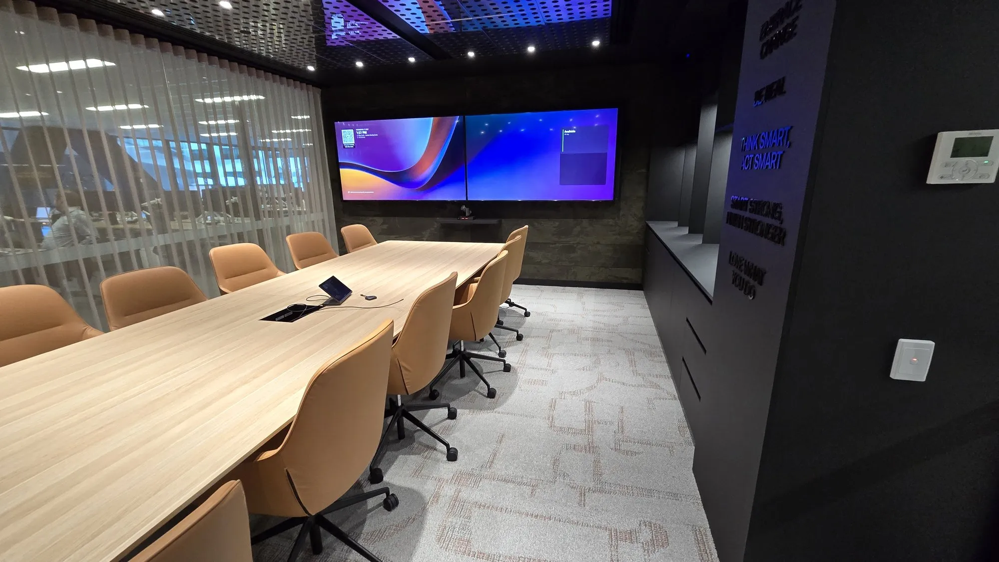 A boardroom with a long timber table, tan leather chairs and dual displays on a dark feature wall