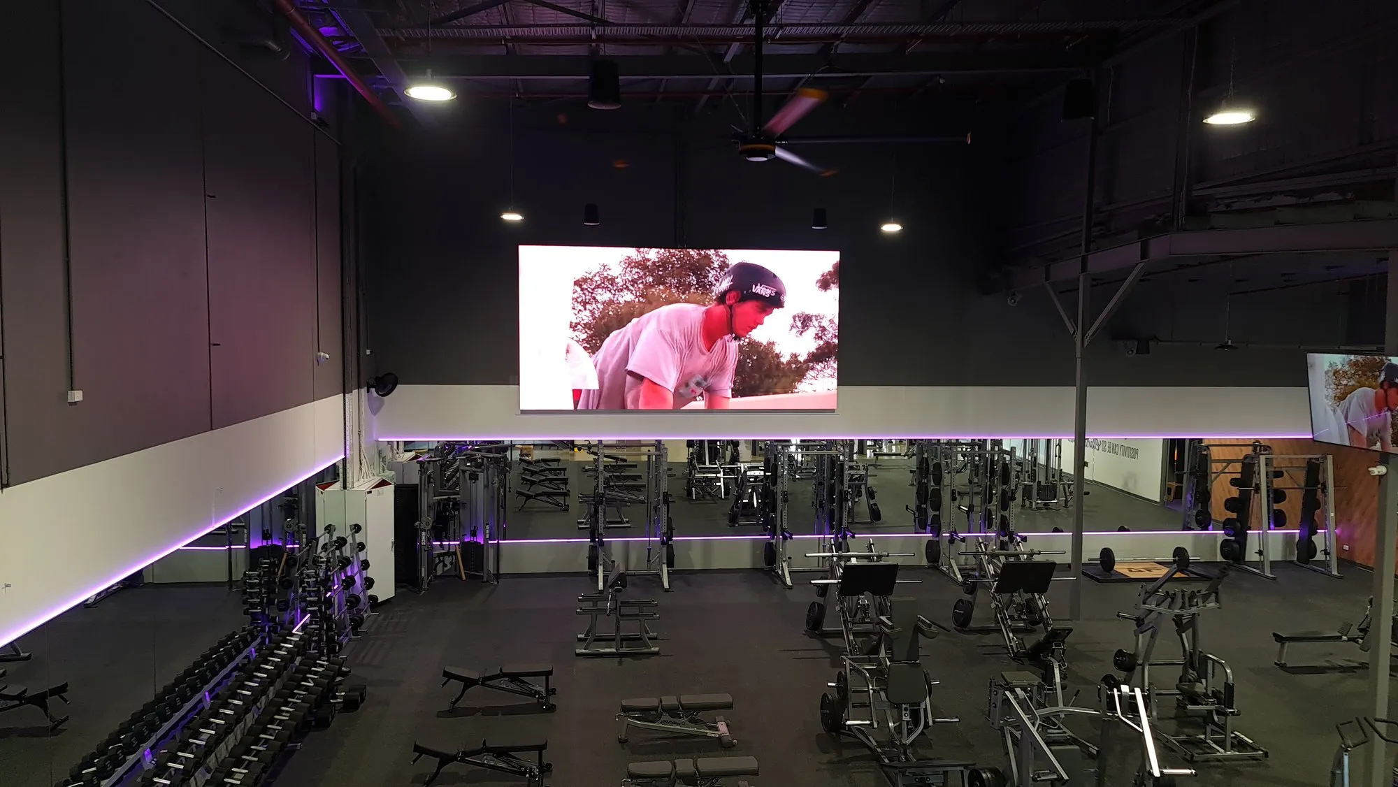 An indoor gym LED wall running rider content above the equipment floor under purple house lighting