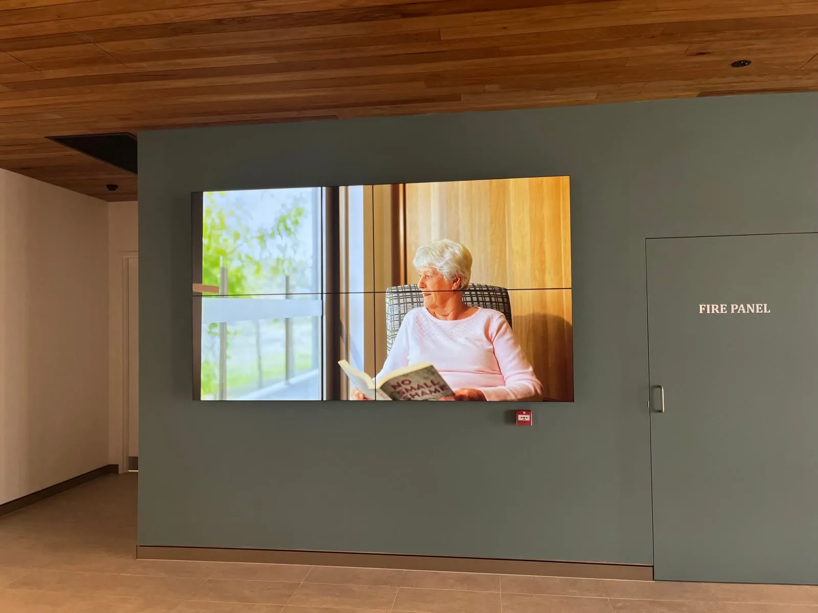A 2×2 video wall on a sage-green wall beneath a timber ceiling at Brightwater Care Group, playing community content
