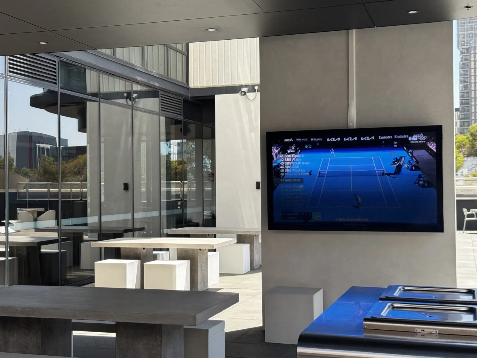 An outdoor terrace screen on a rendered pillar running live tennis with the channel list overlaid, concrete tables and barbecues alongside