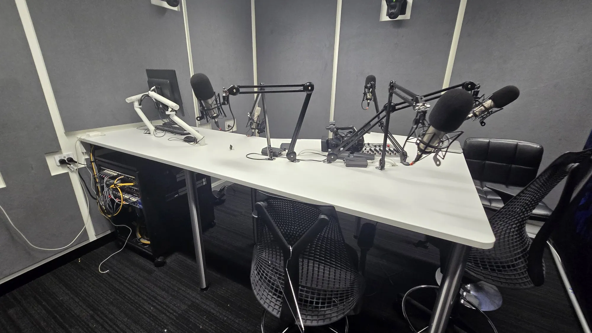 A treated podcast studio — fabric-wrapped panels on the walls and four broadcast microphones on boom arms over the desk