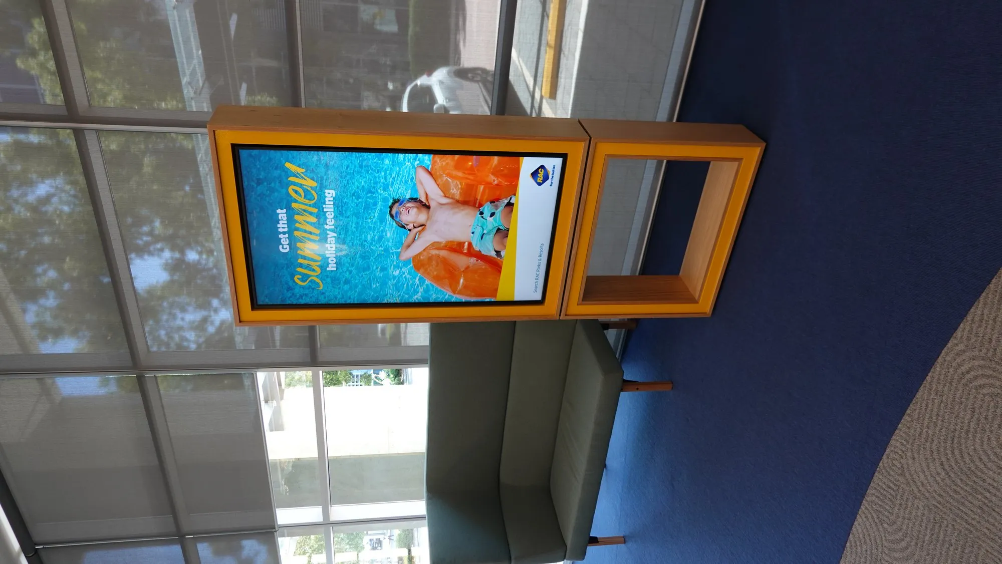 An orange-framed portrait screen angled off the wall, running campaign content at RAC's Perth workplace