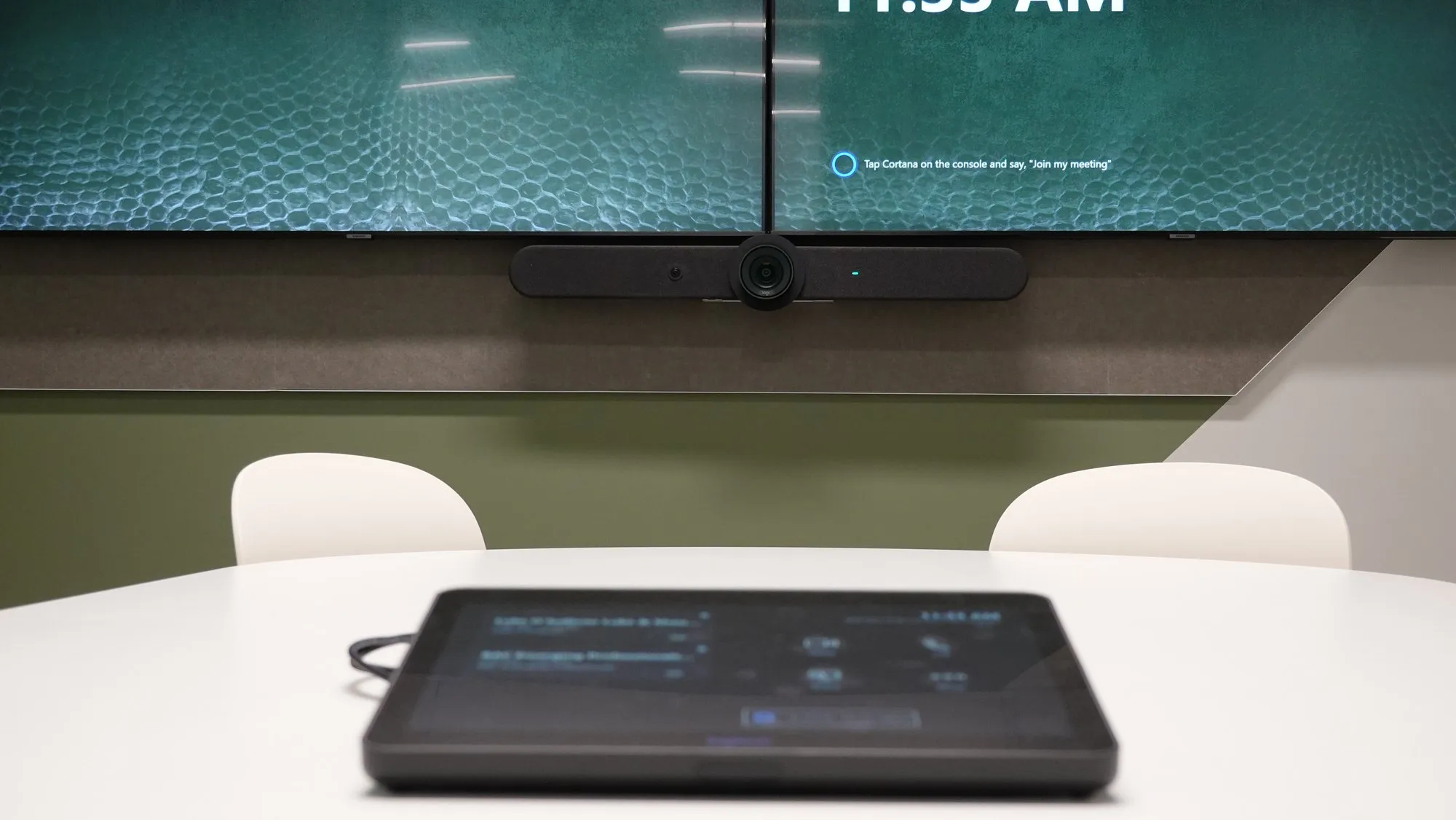 A Logitech Tap console on the boardroom table showing one-tap join, beneath the room's dual displays and video bar