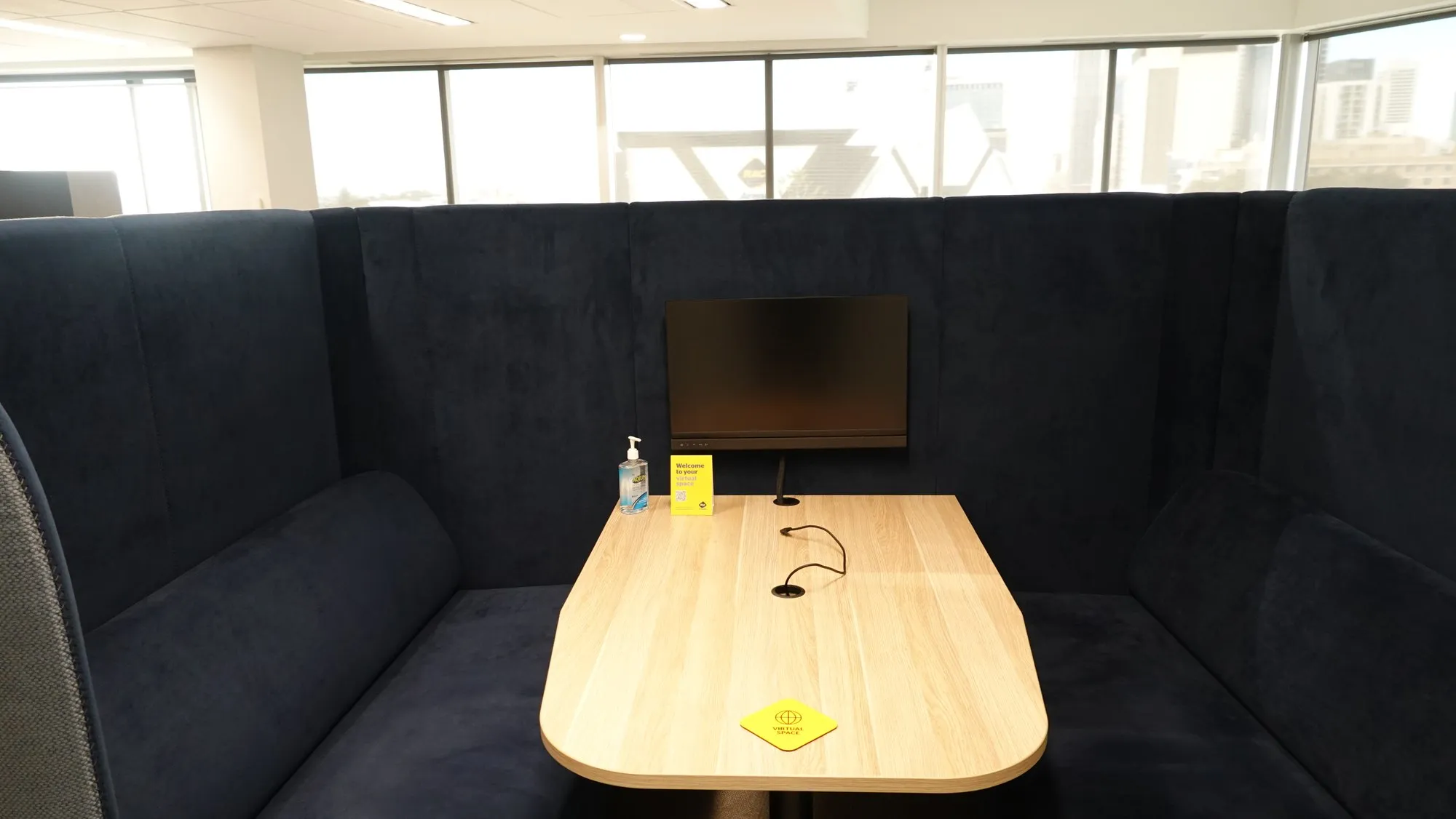 A fabric-walled booth pod with a rounded table and a screen on a low mount, cabled for laptop connection