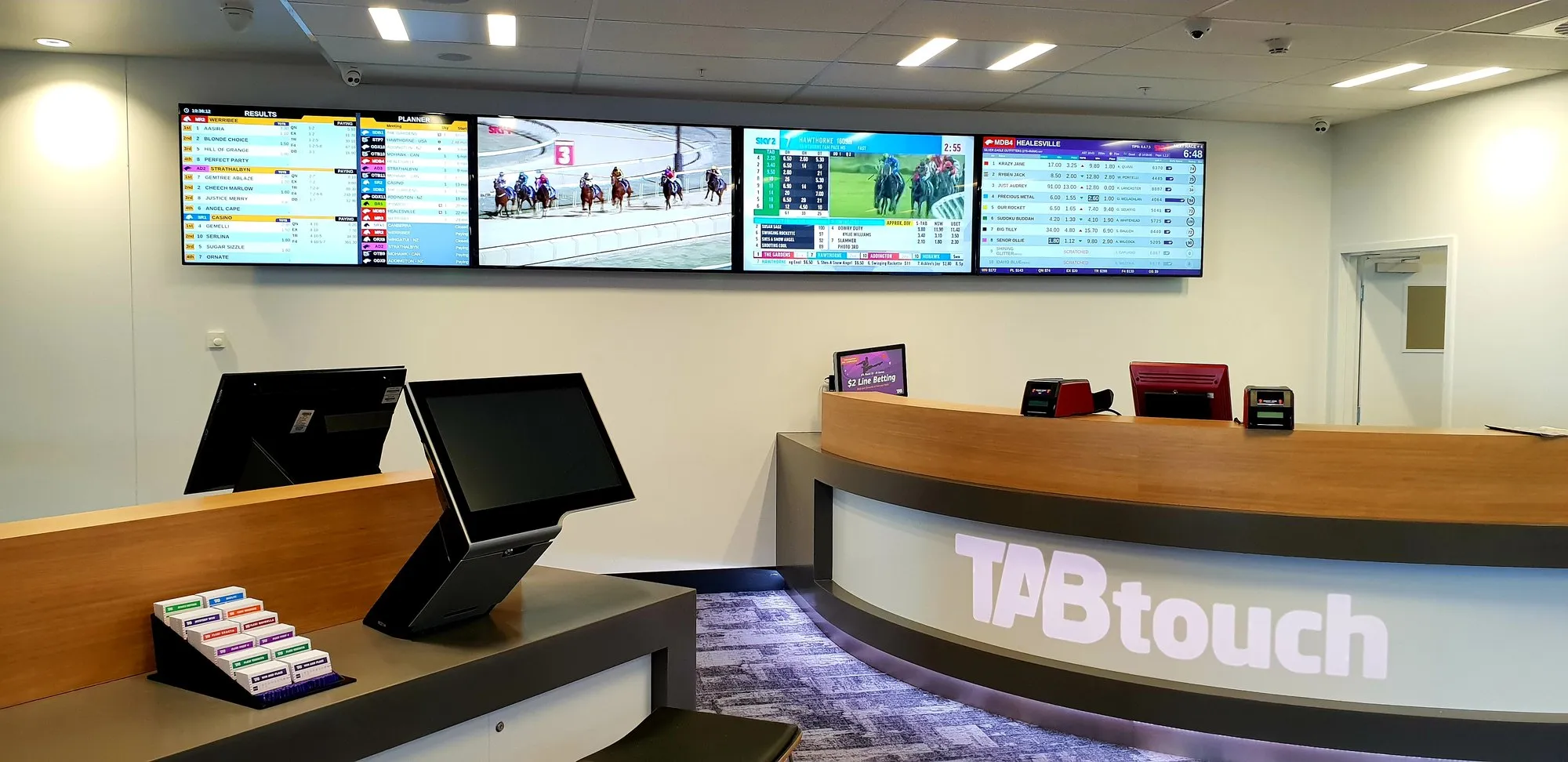 A venue reception with a purpose-built curved counter, service terminals and a bank of screens above it