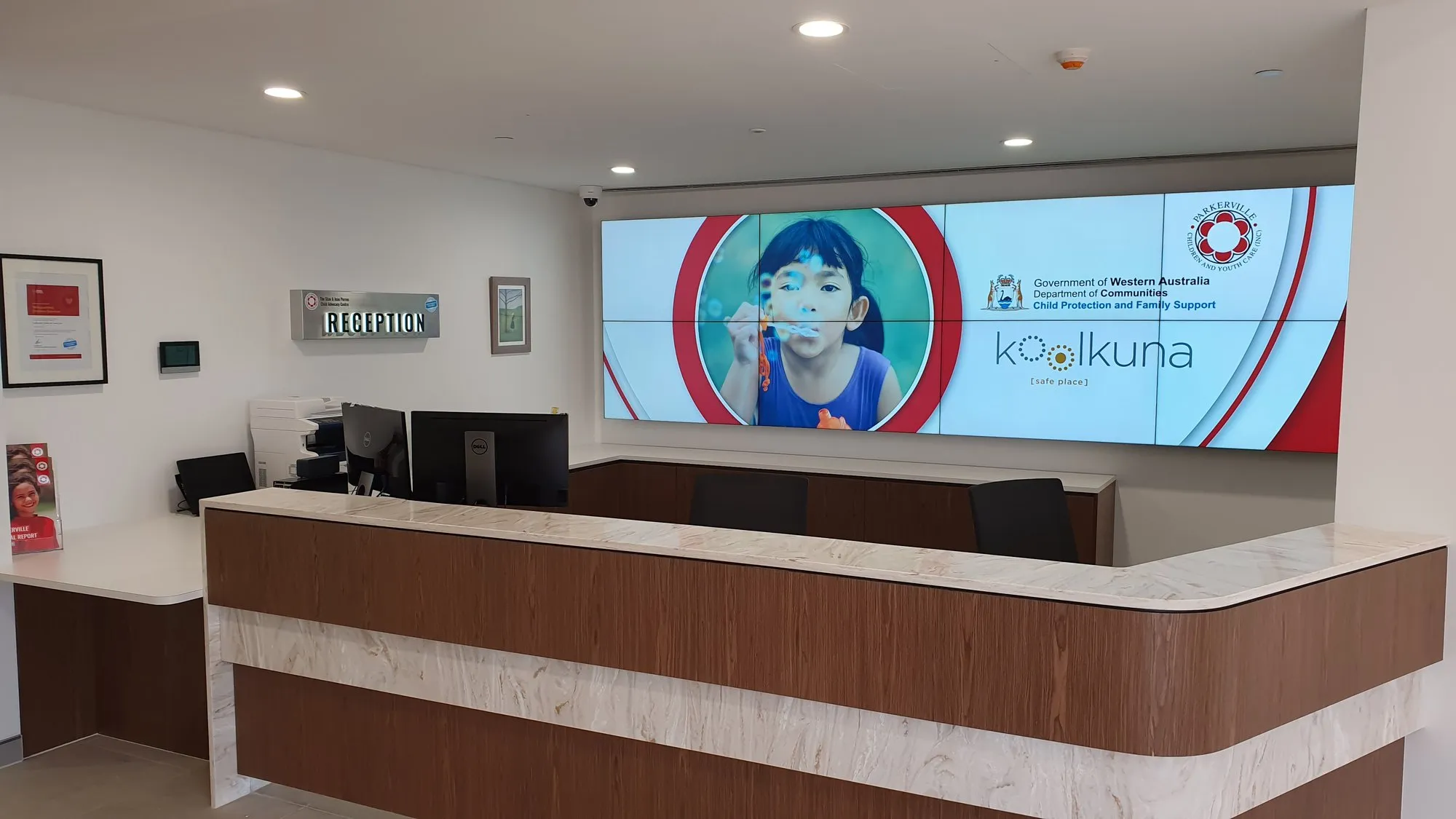 A reception desk backed by a wide LED video wall running a full-bleed campaign image, timber counter in front