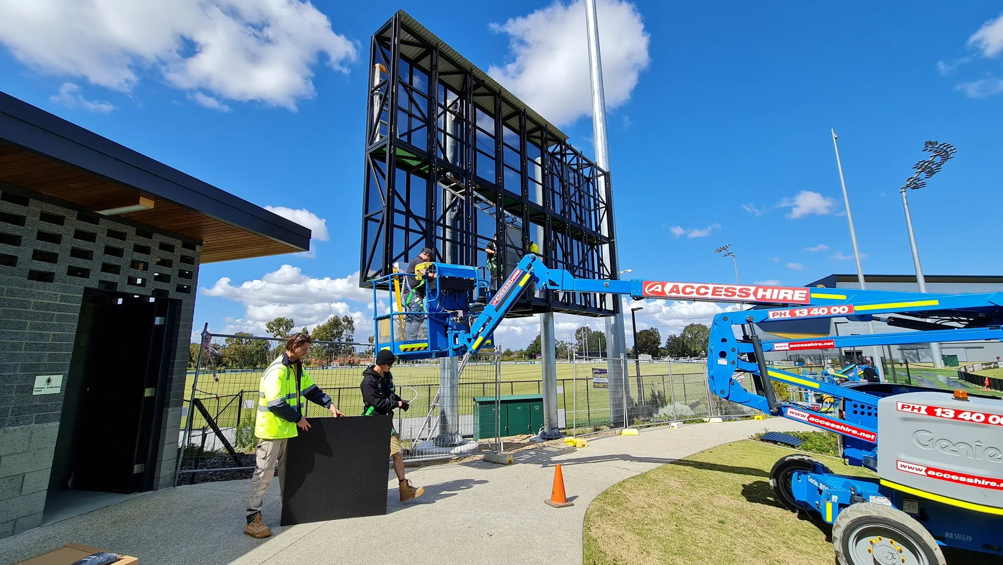 A steel scoreboard frame on poles at a community oval with a boom lift alongside