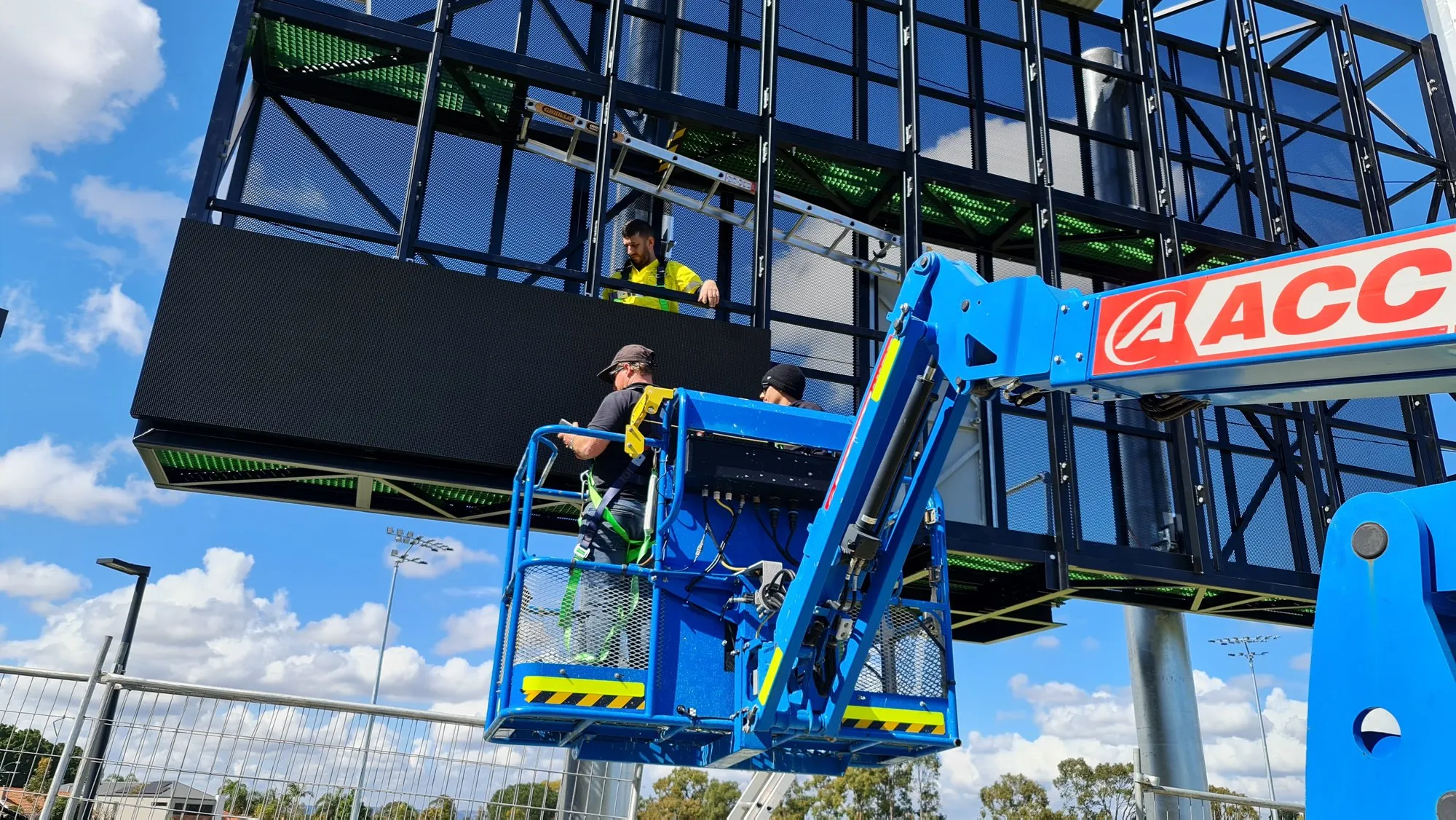 A boom lift raising LED cabinets onto a scoreboard structure against a blue sky
