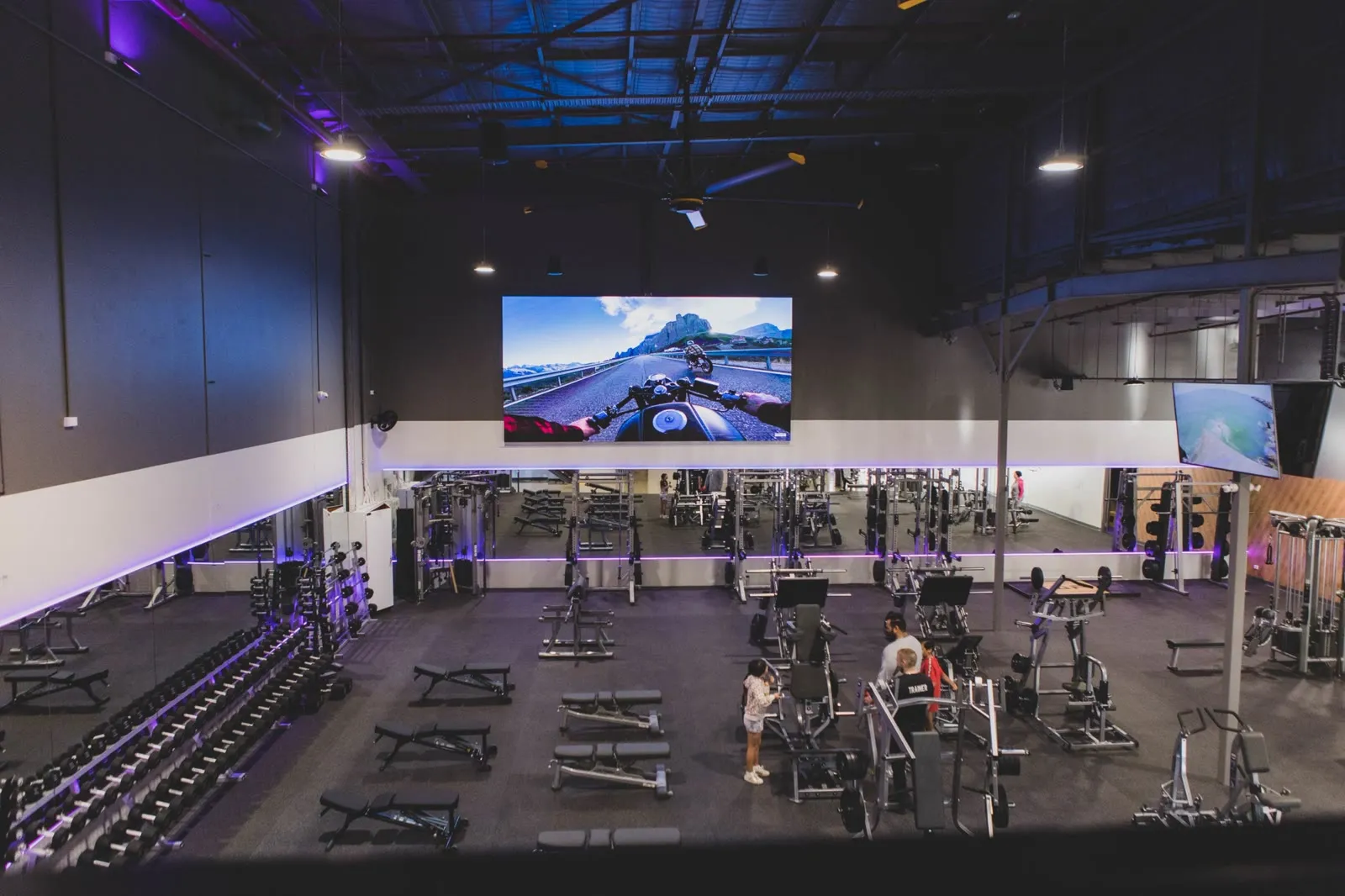 The main gym floor at Anytime Fitness, where a wall-scale screen runs the club's own content channel above the training equipment