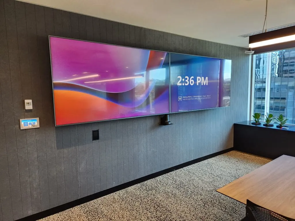 Dual commercial displays mounted on an acoustically treated timber feature wall in CBH Group's Perth boardroom, with a wall control panel beside them and the boardroom table in view
