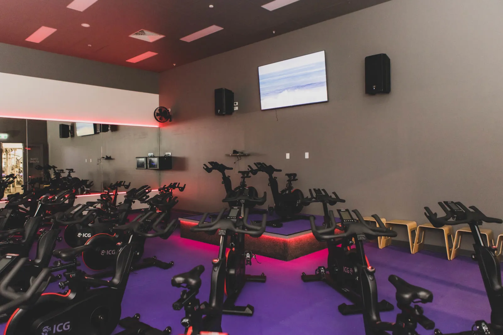 The cycle studio at Anytime Fitness, wall-mounted displays and speakers covering the rows of bikes beneath red ceiling light