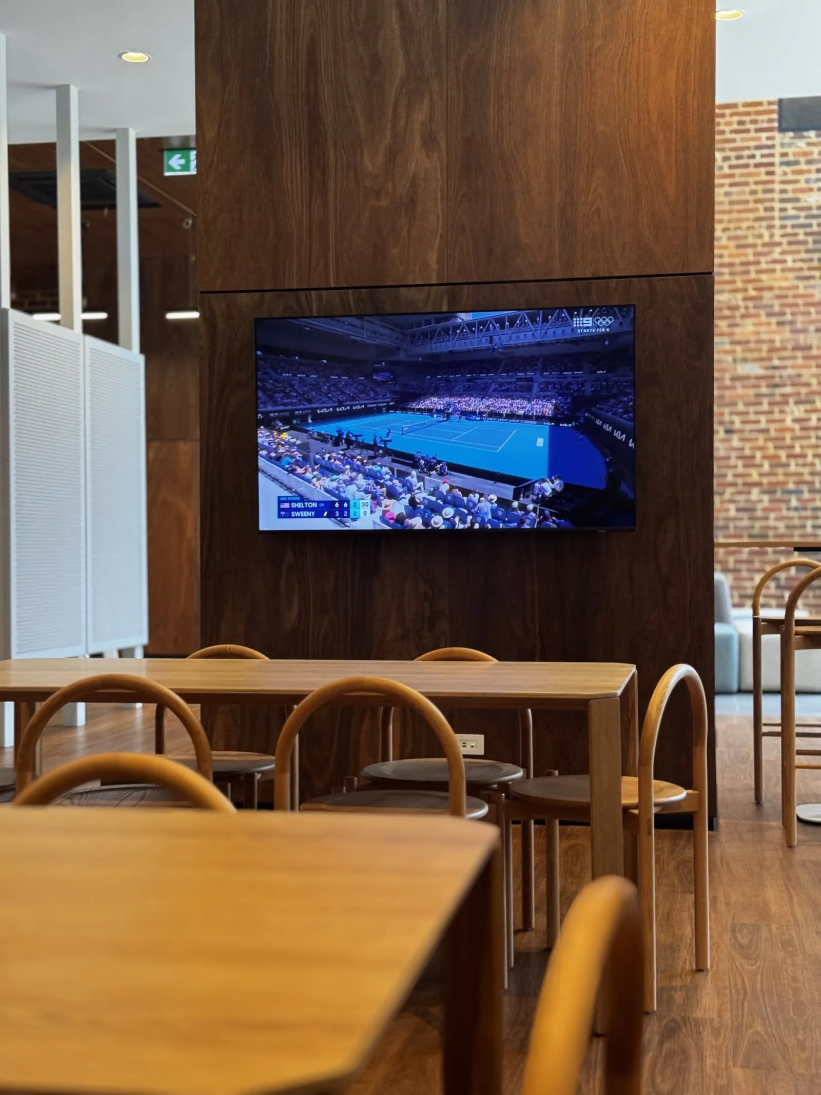 A screen mounted on the timber wall of a workplace café, carrying live sport between the building's content zones