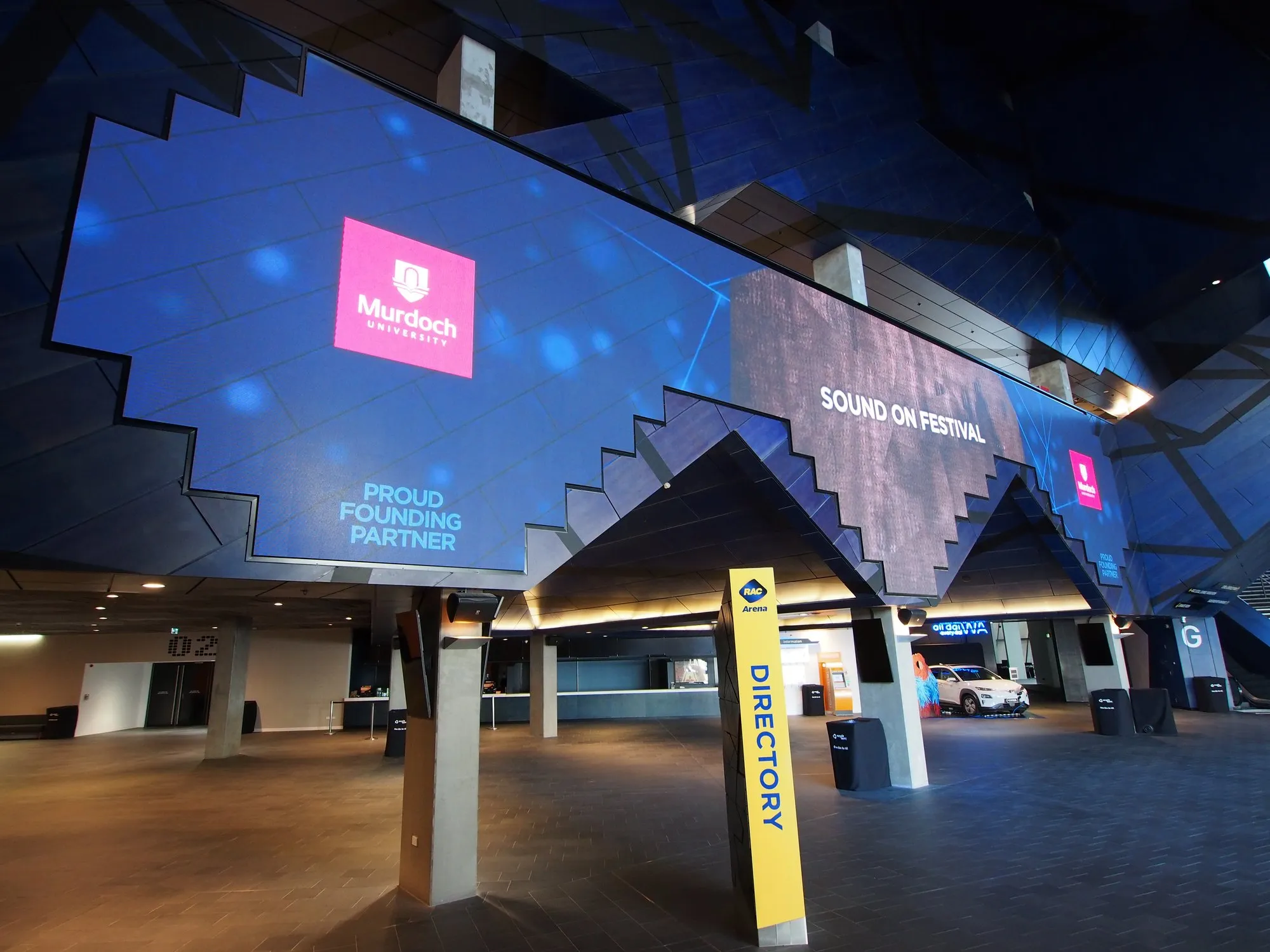 A vast folded LED ribbon arcing across a stadium concourse, lit blue with sponsor and event artwork