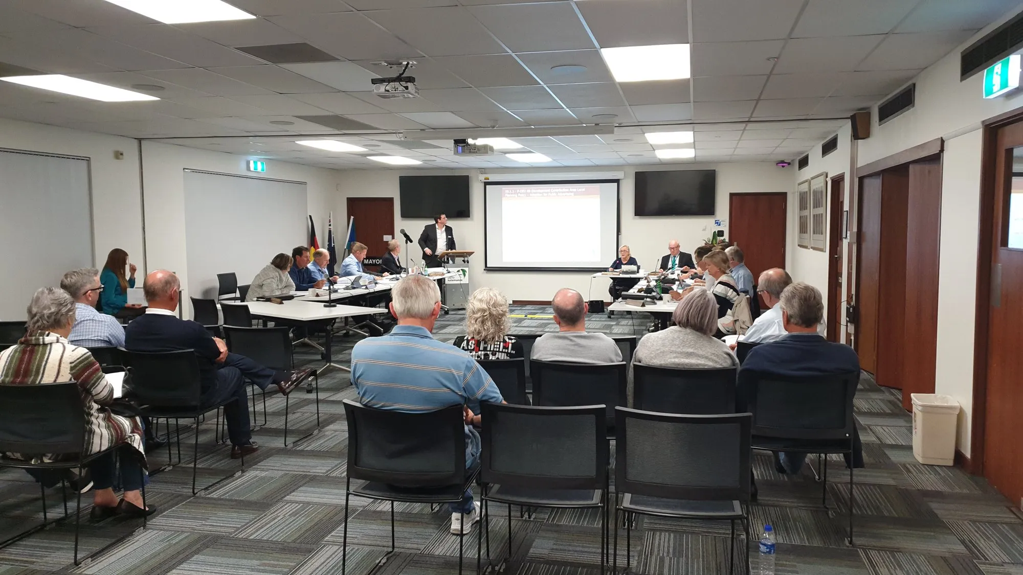 A large training room in session — around twenty-five people seated in rows, presenter at the lectern, projection running