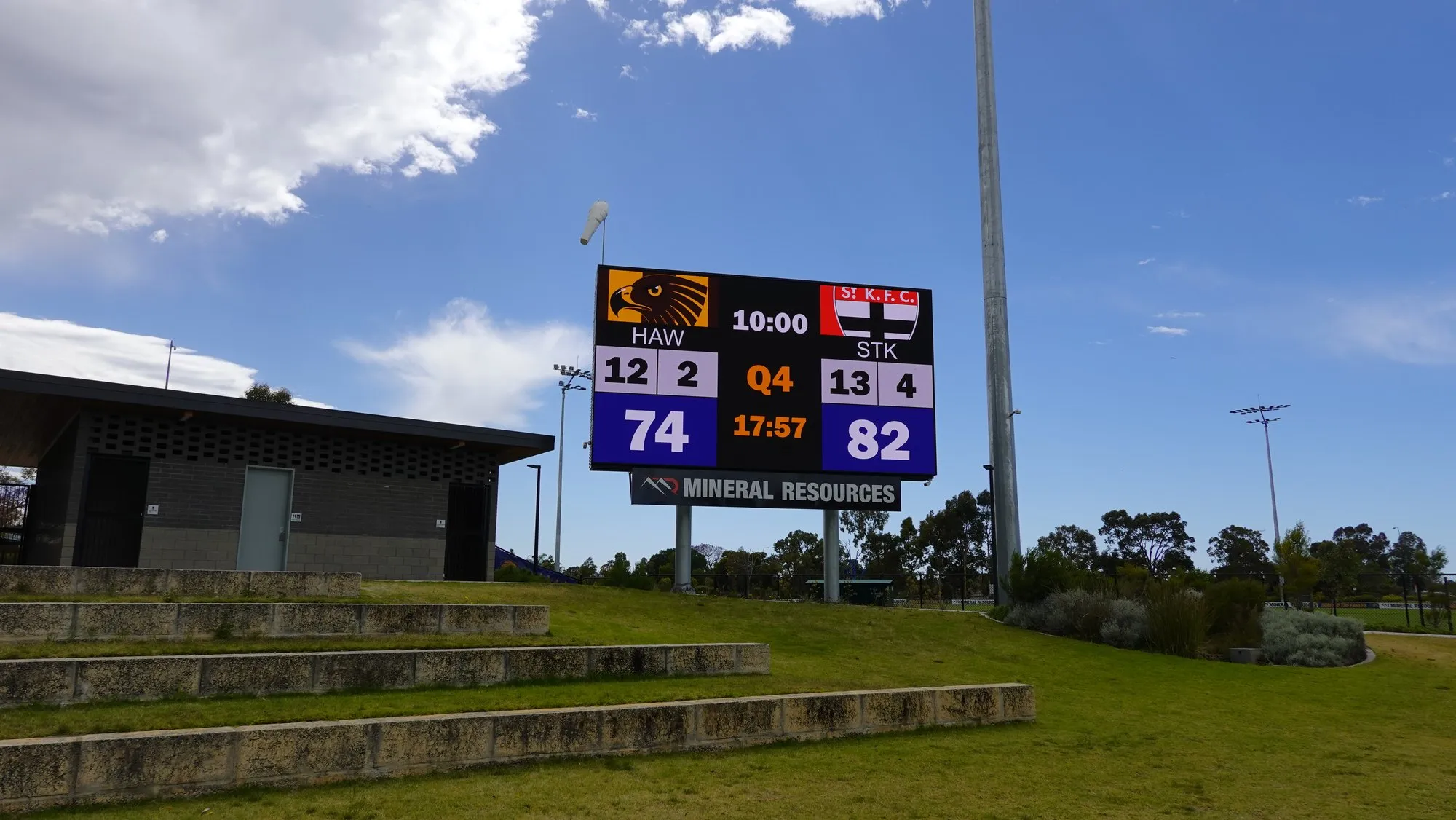 The finished LED scoreboard running a live match score beside terraced seating on a clear day